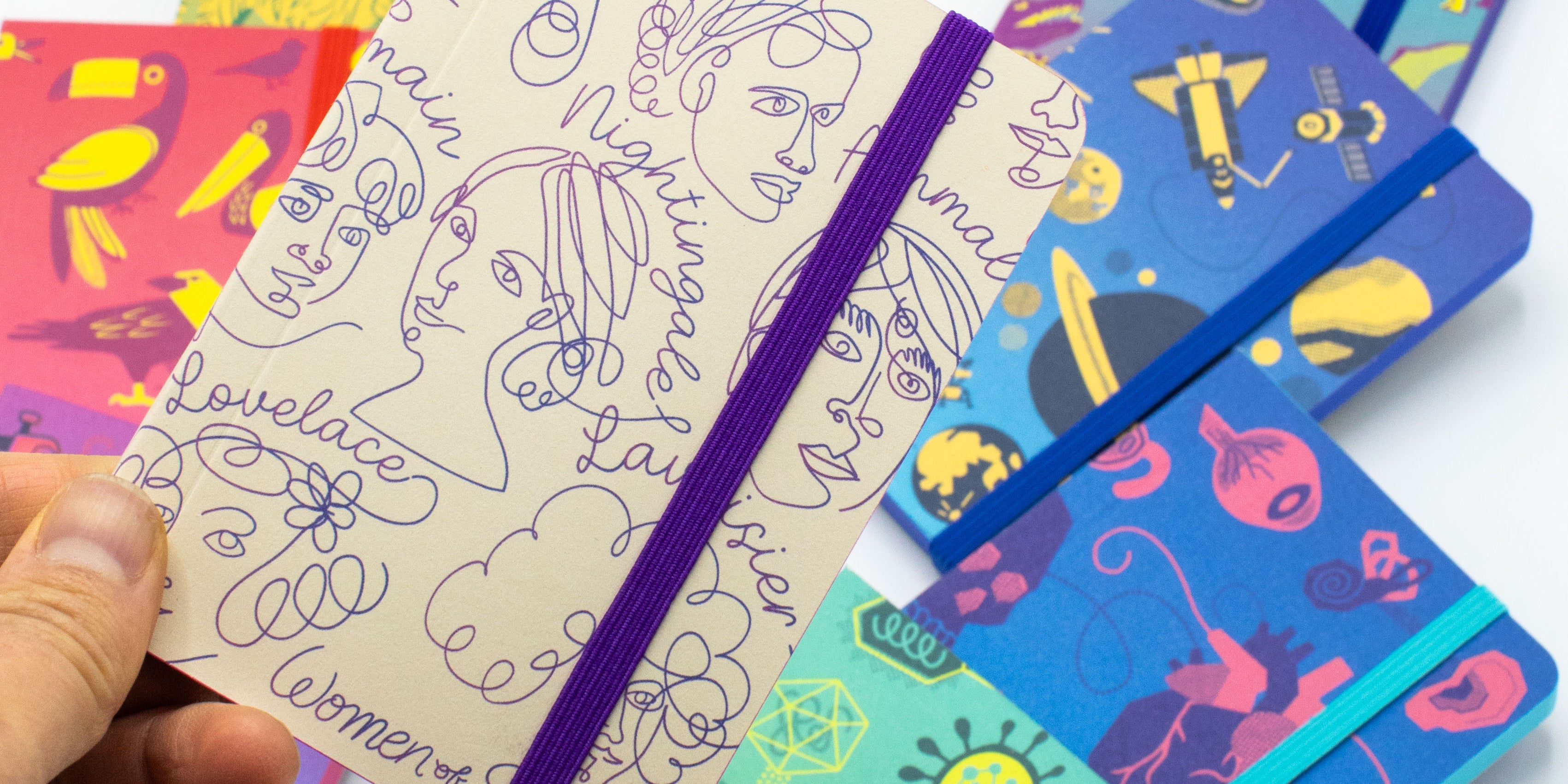 A selection of colorful notebooks laid out on a white surface. A hand holds one notebook featuring abstract line drawings of faces and names.