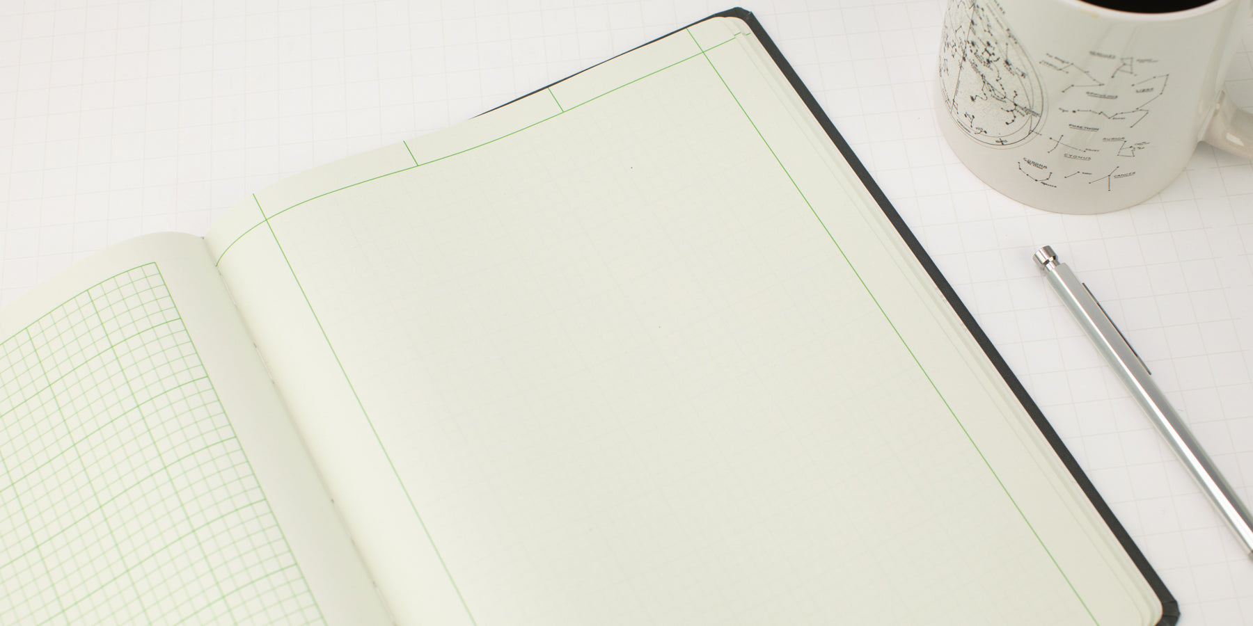 An open notebook with grid paper and blank pages lies on a white surface next to a metal pen and a coffee mug filled with black coffee.