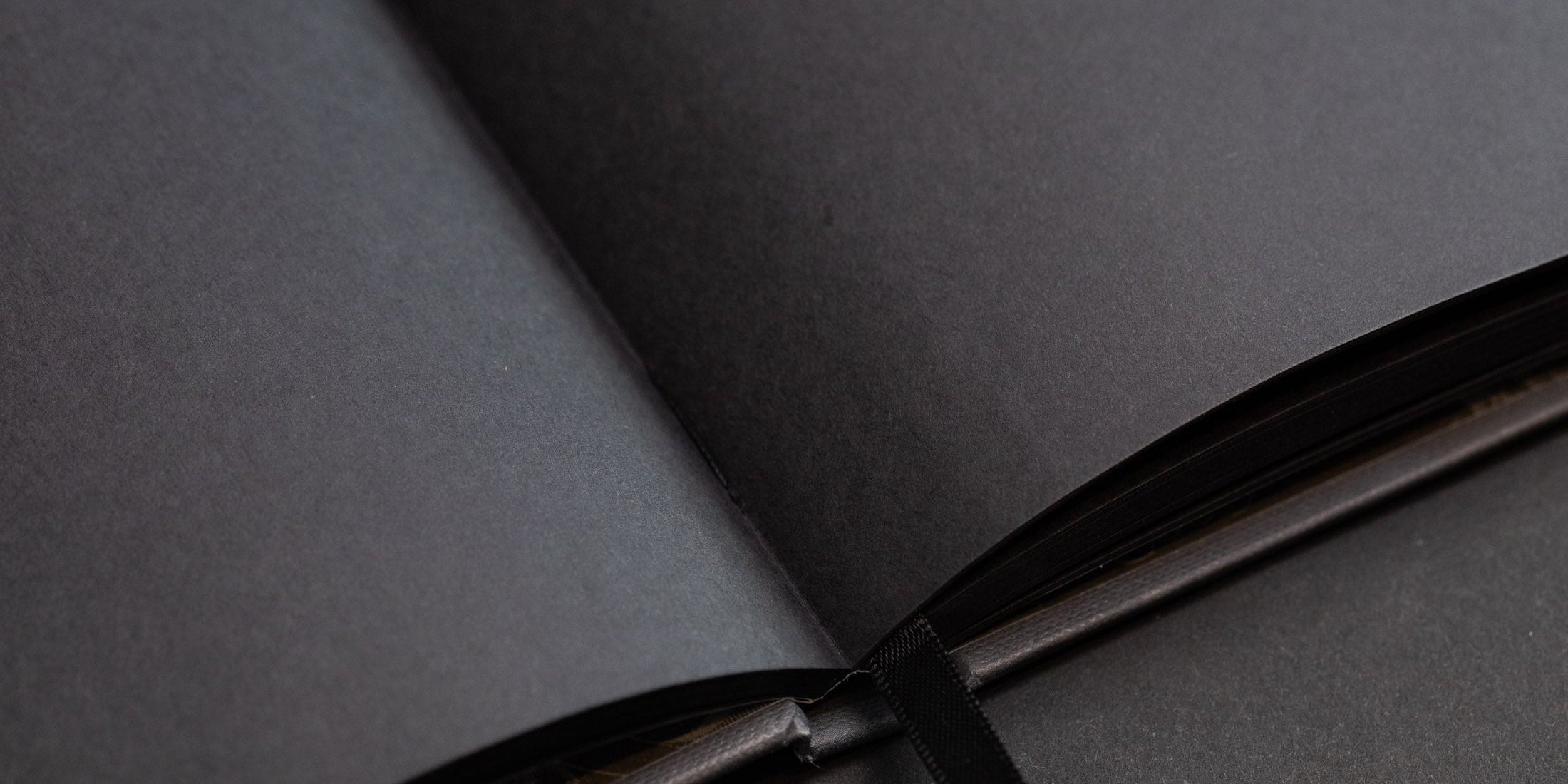 Close-up of an open black book with thick, blank, black pages. A black ribbon bookmark is placed at the center.