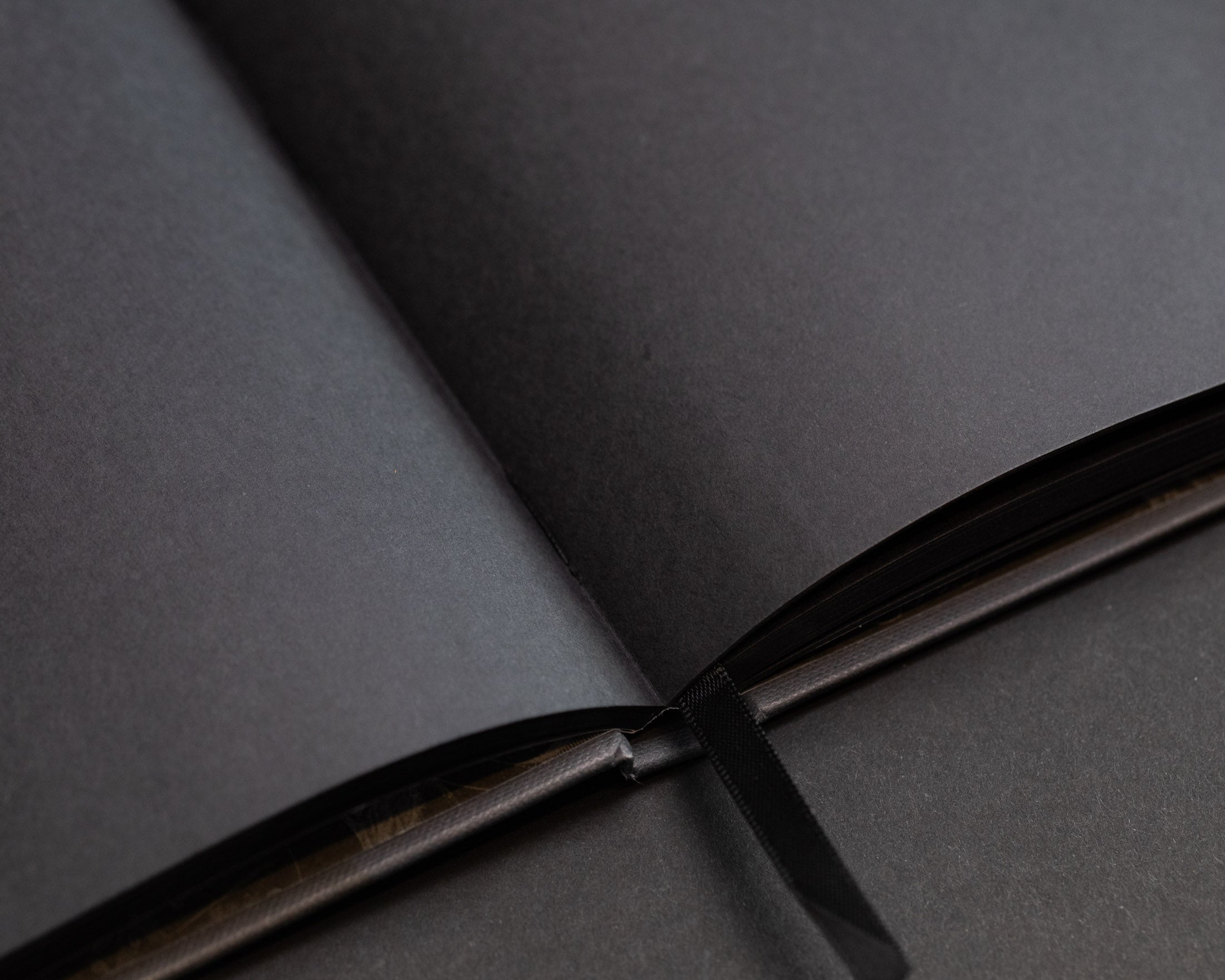 Close-up of an open black book with thick, blank, black pages. A black ribbon bookmark is placed at the center.