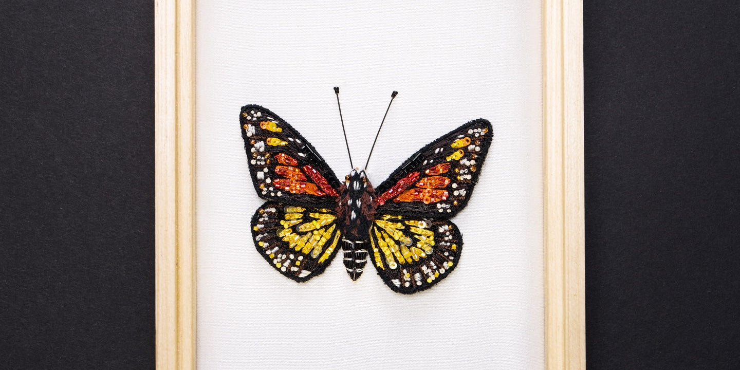 A framed embroidered butterfly with orange, yellow, black, and white wings displayed against a white background on a black surface.