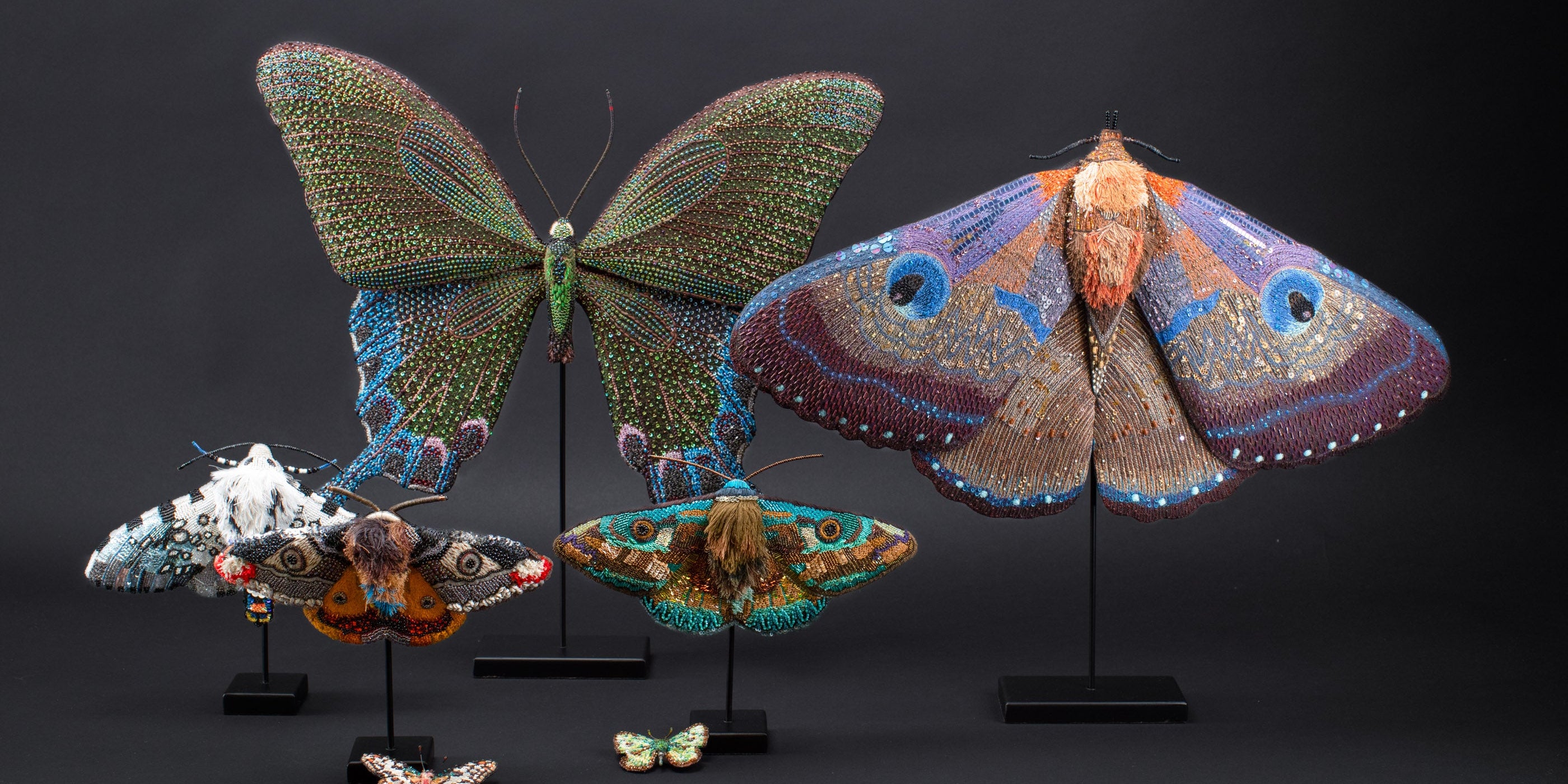 Several intricately beaded sculptures resembling butterflies and moths are displayed on stands against a plain black background.