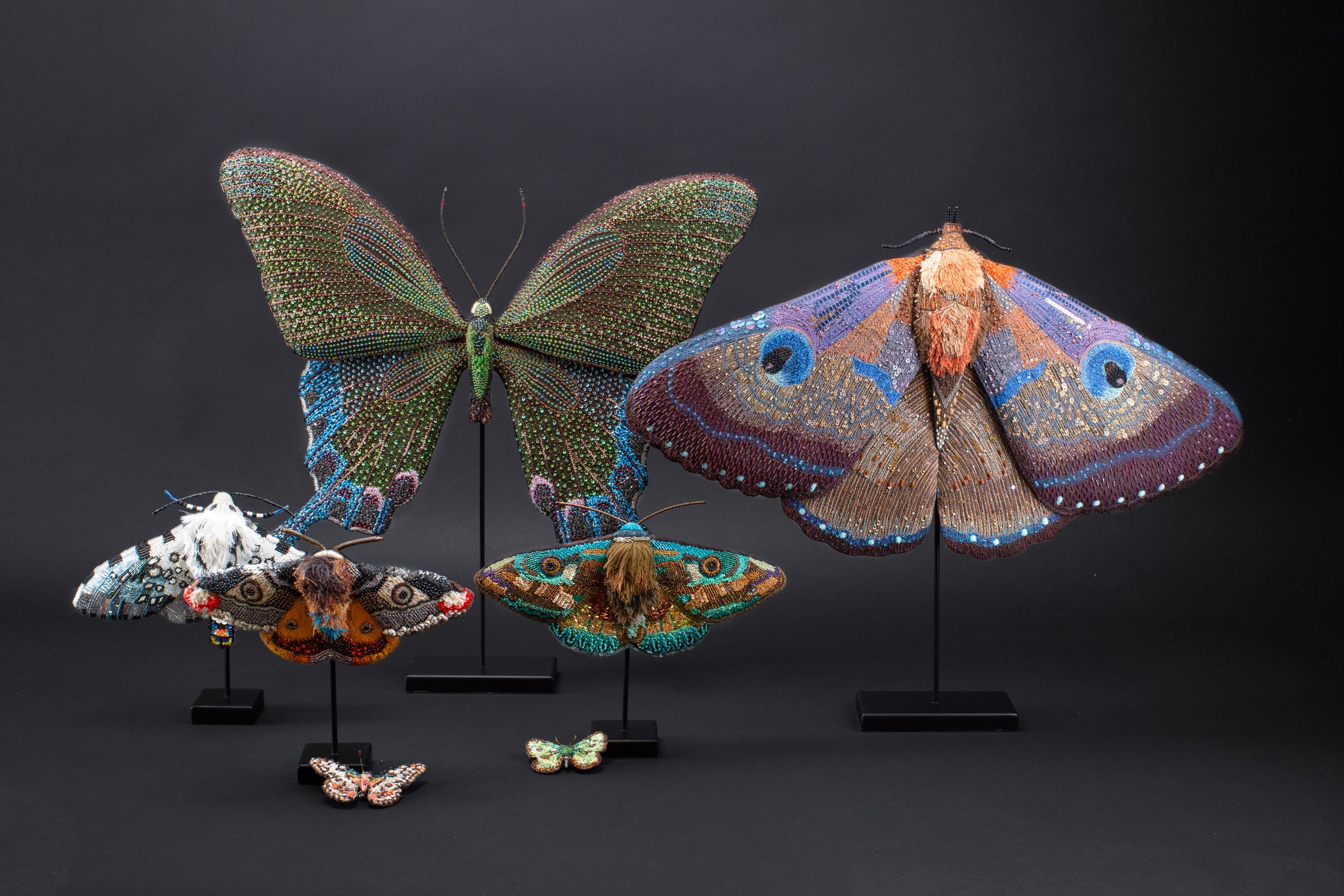 Several intricately beaded sculptures resembling butterflies and moths are displayed on stands against a plain black background.