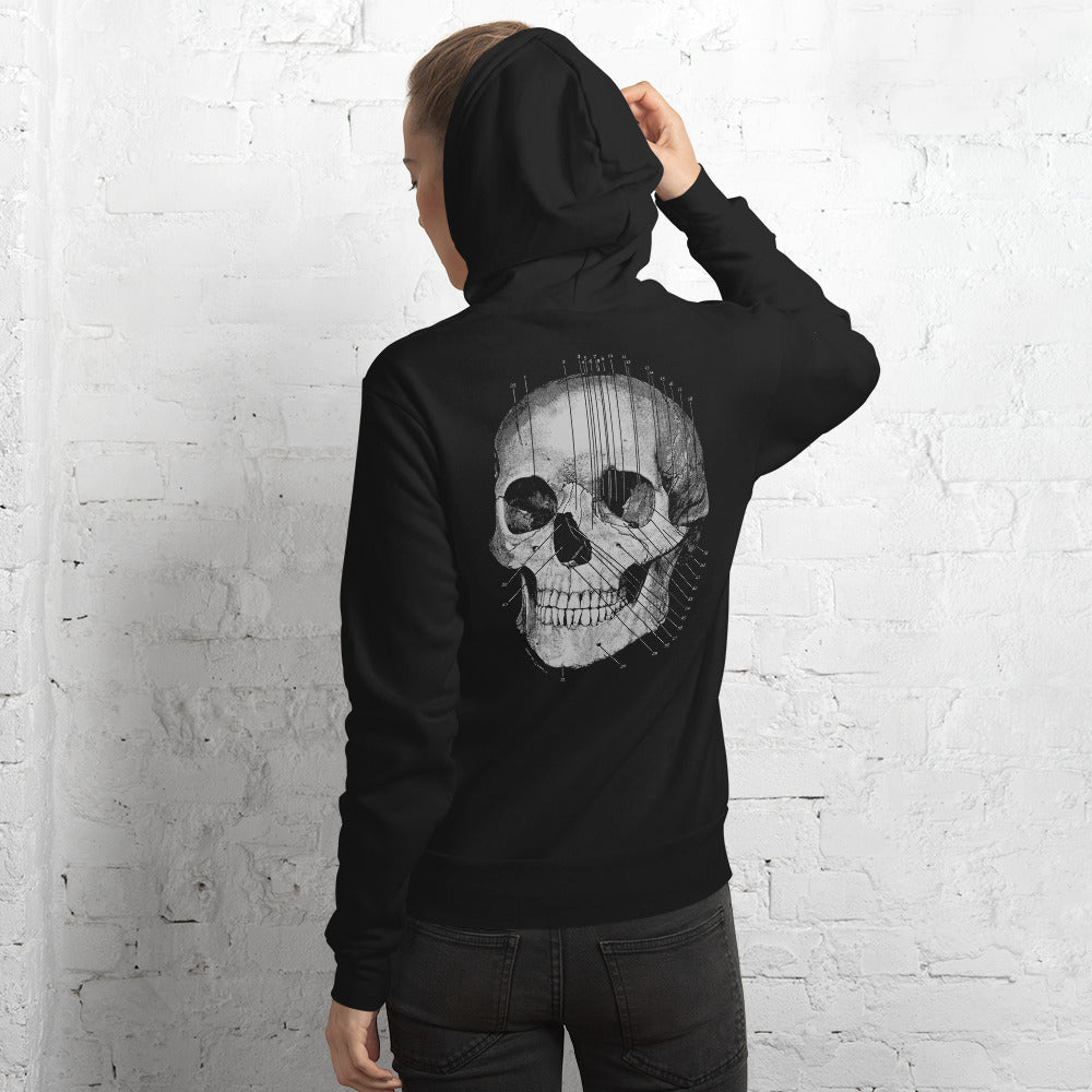 Person wearing a black hoodie with a large skull design on the back stands against a white brick wall.