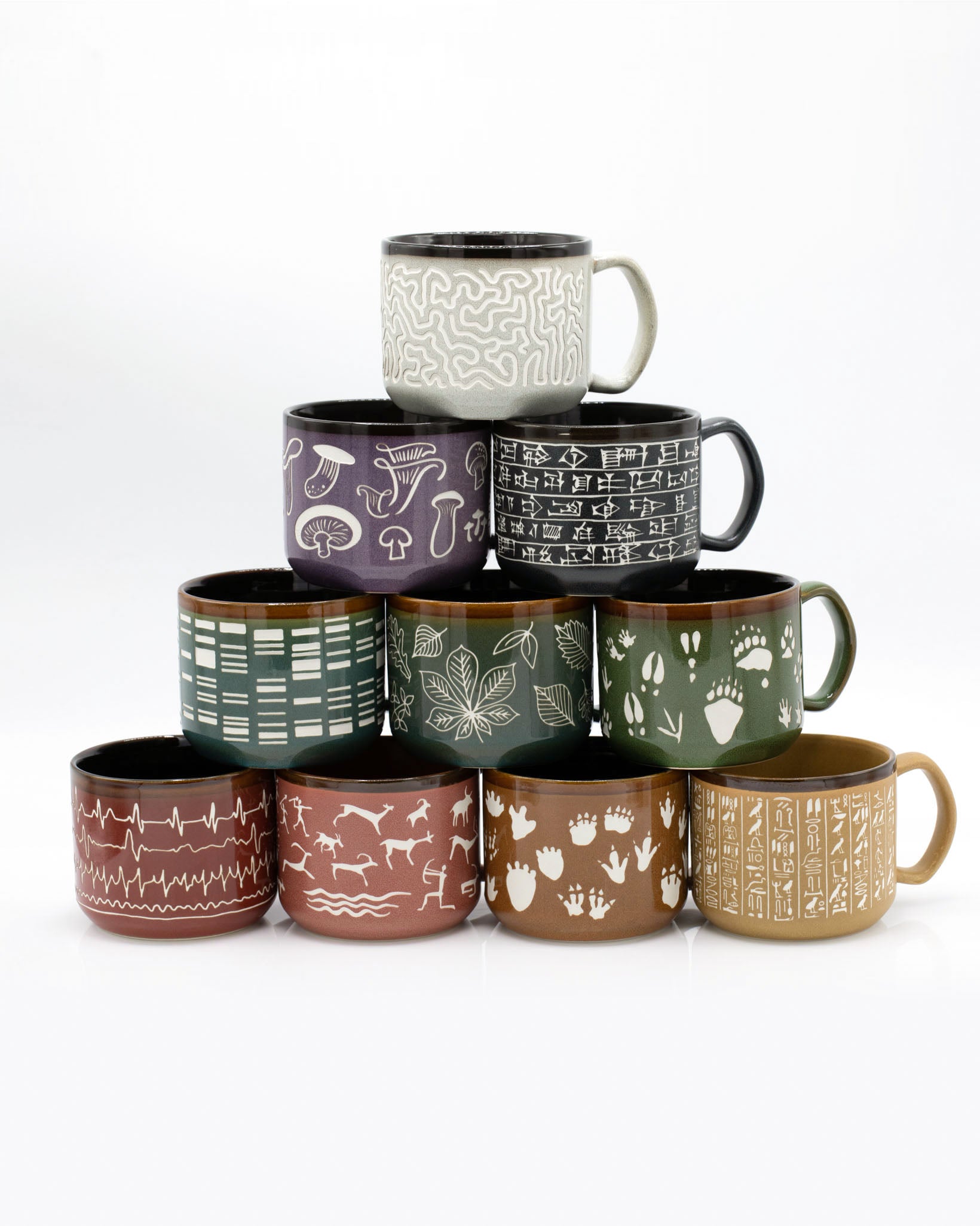 A stack of Hand Carved 15 oz Ceramic Mugs by Cognitive Surplus with different designs on them.